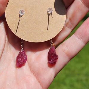 May include: A pair of silver earrings with red gemstone drops. The earrings are on a brown card with a white circle in the centre.