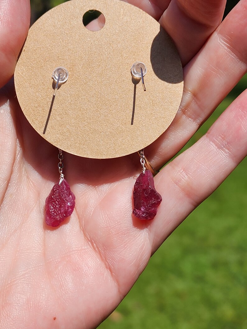 May include: A pair of silver earrings with red gemstone drops. The earrings are on a brown card with a white circle in the centre.