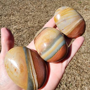 May include: Three polished, oval-shaped stones with a mix of colors including tan, brown, and gray. The stones are held in a hand, showcasing their smooth, layered appearance. The stones have a natural, earthy aesthetic.