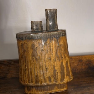 May include: A ceramic vase with a textured, amber-brown body and a dark, glossy grey glaze dripping from the top. The vessel features two distinct cylindrical necks of different heights extending from the flat, dark-glazed upper surface.