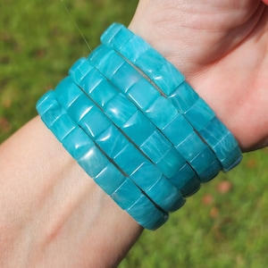 May include: A stack of turquoise blue bracelets made of square-shaped beads. The bracelets are stacked on a person's wrist.