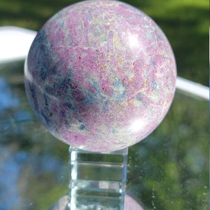 May include: A polished pink and blue gemstone sphere on a clear glass stand. The sphere has a swirling pattern of pink, blue, and white.