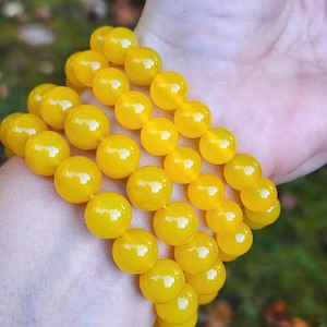May include: A stack of bright yellow, round beads. The beads are strung together, forming a bracelet. The beads are smooth and shiny, reflecting light. The background is blurred, with hints of green and brown.