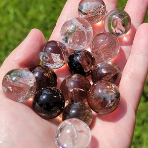 May include: A collection of 12 clear quartz and smoky quartz crystal spheres. The spheres are various sizes and are arranged in a hand.