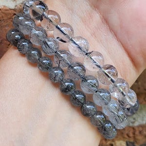 May include: Three layered bracelets made of clear quartz beads with black tourmaline inclusions. The bracelets are stacked on a person's wrist.