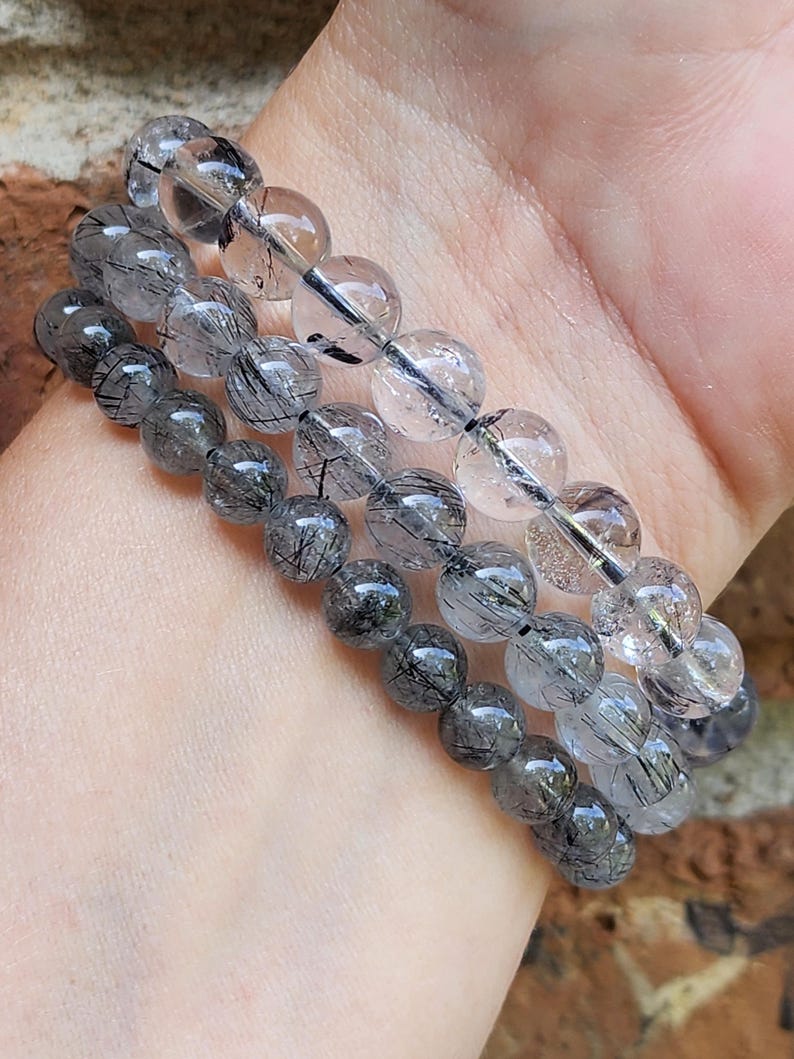May include: Three layered bracelets made of clear quartz beads with black tourmaline inclusions. The bracelets are stacked on a person's wrist.