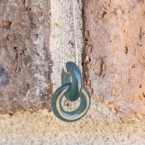 May include: A dark green glass pendant necklace with two interlocking rings. The necklace is hanging from a silver chain against a textured background.