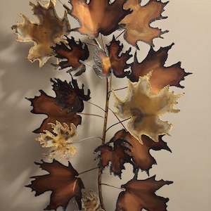 May include: A metal wall art piece featuring a stylized branch with maple leaves. The leaves are in shades of gold, copper, and brown, with dark edges. The artwork is designed to be mounted on a wall, adding a touch of nature-inspired decor.