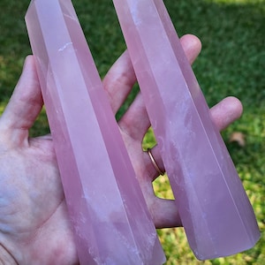 May include: Two pink rose quartz crystal points, also known as wands, are held in a hand. The points are smooth and polished, with a light pink colour.