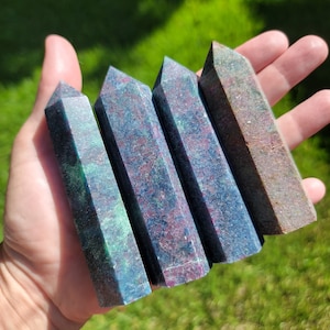 May include: Four gemstone obelisks in shades of blue, green, and red are held in a hand against a blurred green background. The obelisks have a rectangular shape with a pointed top. The stones have a speckled appearance.