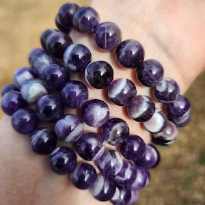 May include: Four amethyst bracelets with white stripes stacked on a person's wrist. The bracelets are made of round beads.