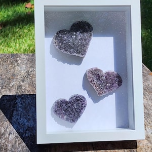 May include: A white framed shadow box containing three heart-shaped amethyst geodes. The geodes are a mix of purple and gray hues, with a sparkling, crystalline texture. The frame is rectangular and sits on a wooden surface.