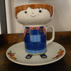May include: A ceramic set consisting of a bowl, a mug, and a plate. The bowl features a cartoon face with brown hair and freckles. The mug depicts a torso in blue overalls and a red plaid shirt. The plate shows a pair of shoes and floral accents.