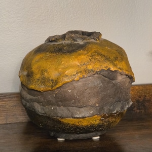 May include: A round, hand-built ceramic vessel with a textured, uneven surface. The piece features a layered aesthetic with a dark, charcoal-gray base and a vibrant, mustard-yellow glaze applied to the upper section and rim, creating a contrast.