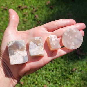 Puede incluir: Cuatro piedras de ágata pulidas de diferentes formas y tamaños. Las piedras son de color rosa claro con marcas blancas y grises. Las piedras están dispuestas en una fila sobre una mano.