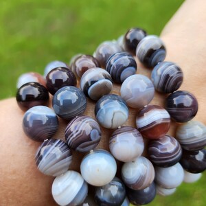 May include: A bracelet made of large, round beads in shades of grey, brown, and white. The beads have a striped pattern and are strung together. The bracelet is worn on a wrist against a blurred green background.