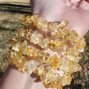 May include: A bracelet composed of multiple strands of polished, golden-yellow citrine gemstone chips. The bracelet is worn on a wrist, with the natural sunlight highlighting the translucent quality of the stones. The bracelet is a piece of jewellery.
