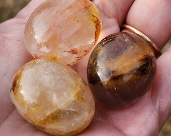May include: Three tumbled stones, one clear with red and orange inclusions, one clear with yellow inclusions, and one brown with gold streaks.