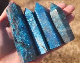 May include: A set of four blue and green apophyllite crystal points, also known as "fairy stones", are arranged in a row in a hand. The crystals are translucent and have a slight shimmer.