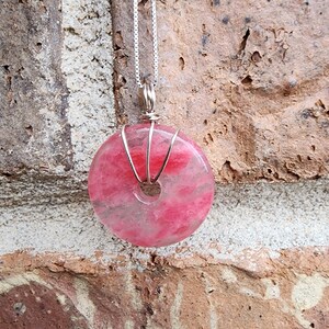 May include: A pink and white donut-shaped pendant necklace. The pendant is made of a translucent stone, wrapped in silver wire, and suspended from a delicate silver chain. The stone has a hole in the center.