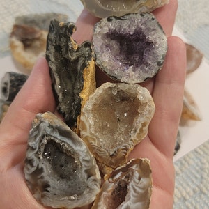 May include: A collection of seven agate geode crystals with various colors and patterns. The geodes are cut open to reveal the crystals inside. The crystals are in various shades of white, gray, brown, and purple.