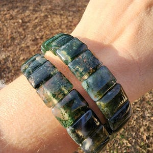 May include: Two moss agate bracelets. Each bracelet is composed of square, polished beads in shades of green, brown, and white. The bracelets are worn on a wrist, showcasing the natural patterns and colors of the gemstones.