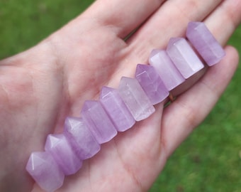 May include: A collection of ten small, polished amethyst points in various sizes, arranged in a row on a hand. The points are a pale lavender color and have a smooth, glassy finish.
