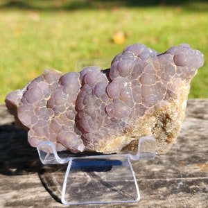 May include: A large, purple, agate geode with a bubbly, textured surface. The geode is displayed on a clear plastic stand.