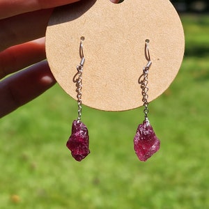 May include: A pair of earrings with a silver chain and a raw, deep red ruby gemstone on each earring.