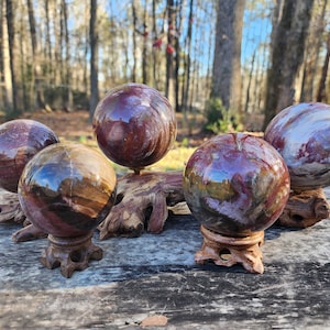 May include: Five polished, colorful, agate spheres on wooden stands. The spheres are a mix of red, brown, and white colors. The spheres are on a wooden surface.