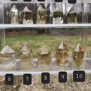 May include: Ten clear quartz crystal points with a golden yellow hue, displayed in a clear acrylic case. The crystals are arranged in two rows of five, with numbers 1 through 10 labeled above each crystal.