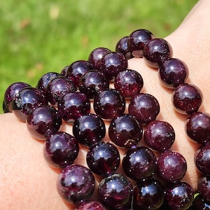 May include: A close-up of a stack of garnet gemstone bracelets. The bracelets are made of round, dark red beads with a shiny finish.