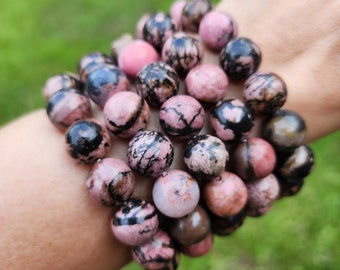 May include: A bracelet made of round, polished beads. The beads are primarily pink with black and brown veining. The bracelet is worn on a wrist, with a blurred green background.