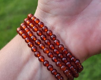 May include: Four strands of reddish-brown beaded bracelets are stacked on a wrist. The beads are small, round, and have a glossy finish. The background is blurred green grass.