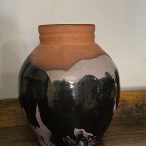 May include: A ceramic vase with a rounded body and a short, straight neck. The upper portion of the vase features an unglazed, matte terracotta finish, while the lower section is coated in a glossy black glaze with white marbled accents.