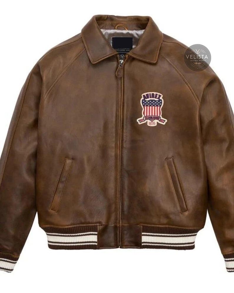 May include: Brown leather jacket with a zip-up front, a collar, and a patch on the chest. The patch features a design with red, white, and blue colors. The jacket has two front pockets and ribbed cuffs and hem with white stripes.