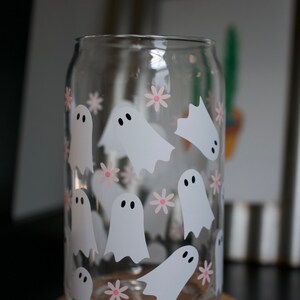 May include: A clear glass can with a design of white ghosts and pink flowers. The can is sitting on a cork coaster.