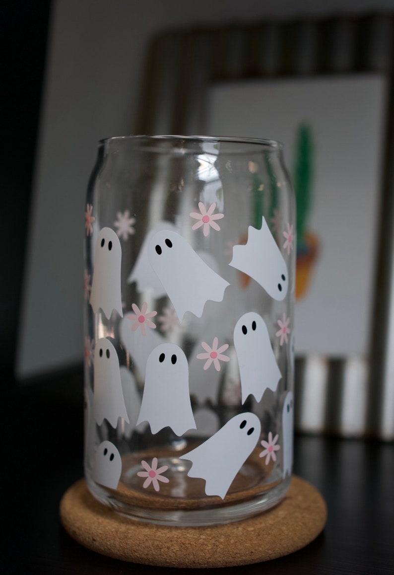 May include: A clear glass can with a design of white ghosts and pink flowers. The can is sitting on a cork coaster.