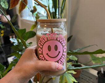 Può includere: Un bicchiere con un coperchio di legno e una cannuccia dorata riempito di una bevanda marrone. Il bicchiere ha un'emoticon sorridente rosa con il testo "Find the good vibes" intorno.