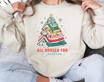 May include: A cream-coloured sweatshirt with a Christmas tree design made of stacked books with colourful lights. The text "All Booked for Christmas" is printed below the tree.
