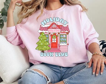 May include: Pink sweatshirt featuring a cartoon illustration of a house with a Christmas tree and the text "Holiday Bookstore Book Club".