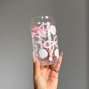 May include: A clear glass can-shaped drinking glass decorated with pink pearl strands, bows, and white seashell graphics. The glass is held up against a gray background.