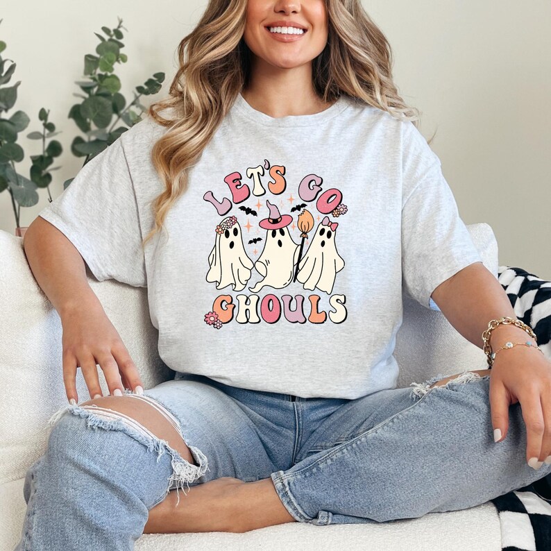 May include: A gray t-shirt with a graphic of three ghosts wearing witch hats and holding a broom. The text "Let's Go Ghouls" is printed in pink and white letters.