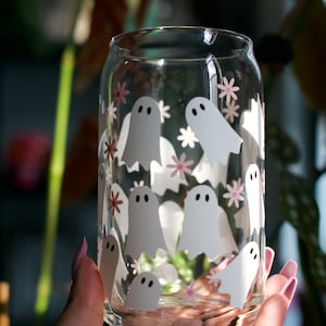 May include: A clear glass can with a white ghost and pink flower pattern. The can is being held by a hand.