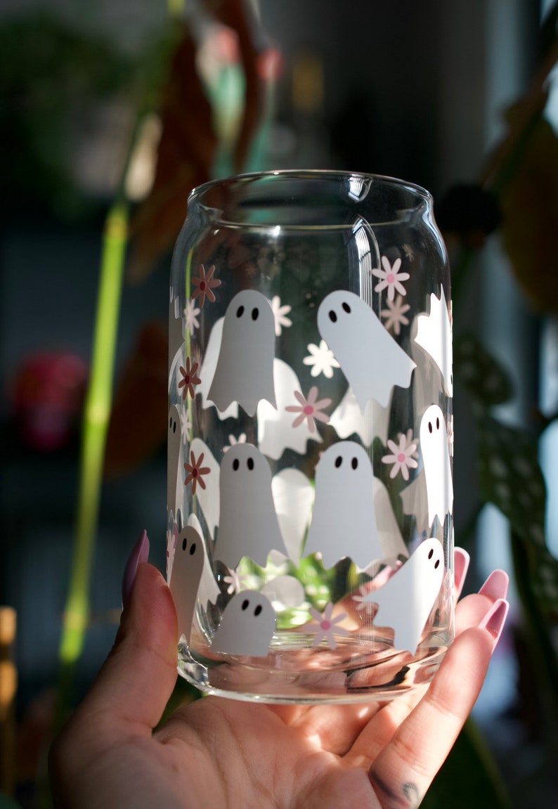 May include: A clear glass can with a white ghost and pink flower pattern. The can is being held by a hand.