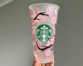 May include: A clear, reusable Starbucks tumbler with a pink cherry blossom design. The Starbucks logo is in green. The cup is held by a hand with a manicure.