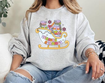 May include: A grey sweatshirt featuring a graphic of a sleigh filled with books and presents. The text "Seasons Readings" is written on the sleigh.