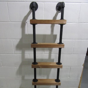 May include: A wall-mounted ladder shelf with an industrial aesthetic. It has four wooden shelves held by black metal pipes and fittings. The ladder is designed for wall mounting, with two black metal brackets at the top. The shelves are approximately 60 cm wide.