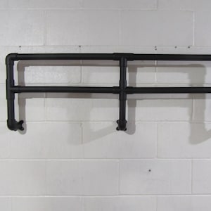 May include: Black metal pipe shelf, mounted on a white brick wall. The shelf is constructed from connected pipes and fittings, creating a two-tiered design. The shelf is designed to be mounted on a wall.