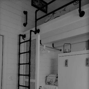 May include: A black metal ladder leading up to a loft bed with a white wooden frame. The loft bed has a white wooden closet with a white door and a shelf above it. The closet has a black metal clothes rail.